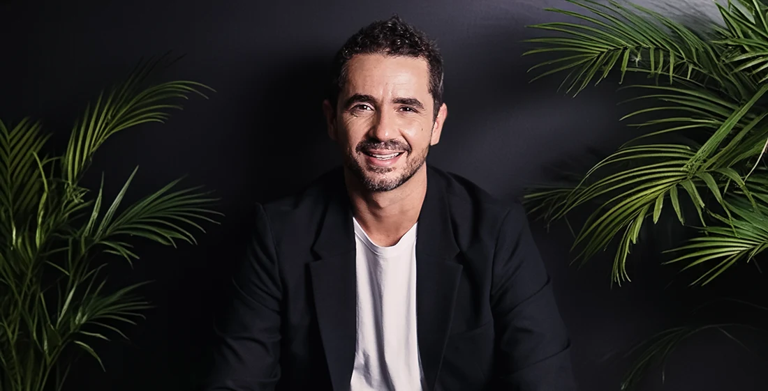 Foto de Felipe Andreoli vestindo blazer preto, camiseta branca. Atrás dele tem uma parede preta com folha de palmeiras nos cantos.